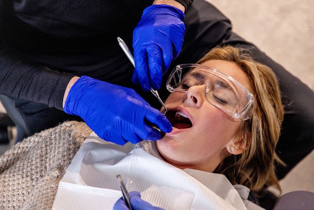 Dentist performing an oral examination on a patient wearing protective eyewear, with gloved hands holding dental instruments.
