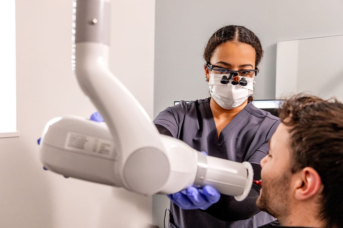 Dental professional wearing protective eyewear and a mask positioning a digital X-ray machine to take dental images of a male patient in a dental clinic.