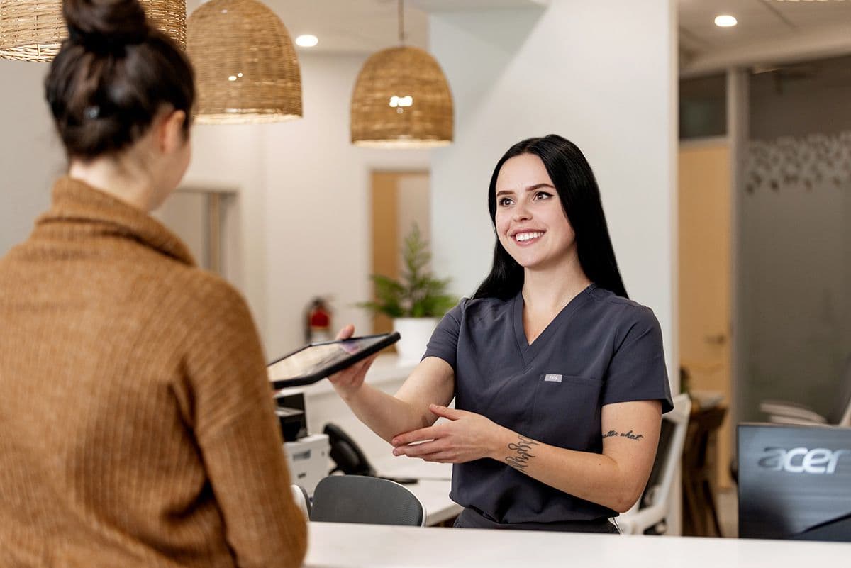 Dental receptionist at Miller Dental Orillia smiling and handing a digital tablet to a patient at the front desk during check-in.