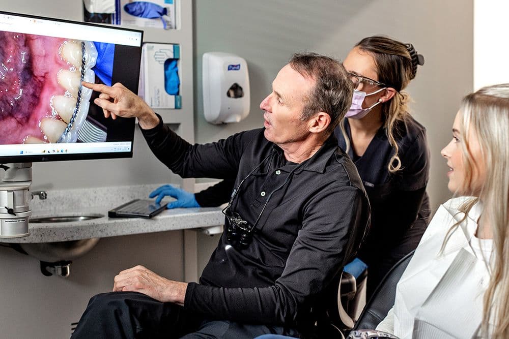 Dentist pointing to a close-up image of teeth with braces on a computer screen while explaining treatment to a patient, with a dental assistant standing nearby in a dental clinic.Dentist pointing to a close-up image of teeth with braces on a computer screen while explaining treatment to a patient, with a dental assistant standing nearby in a dental clinic.