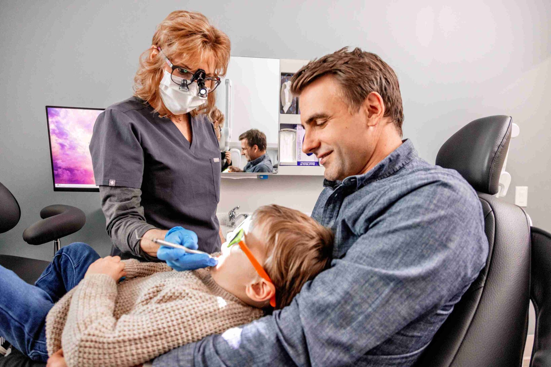 Dentist providing a gentle dental exam for a young child sitting on a parent’s lap, demonstrating family-friendly and accessible dental care at Miller Dental Care.